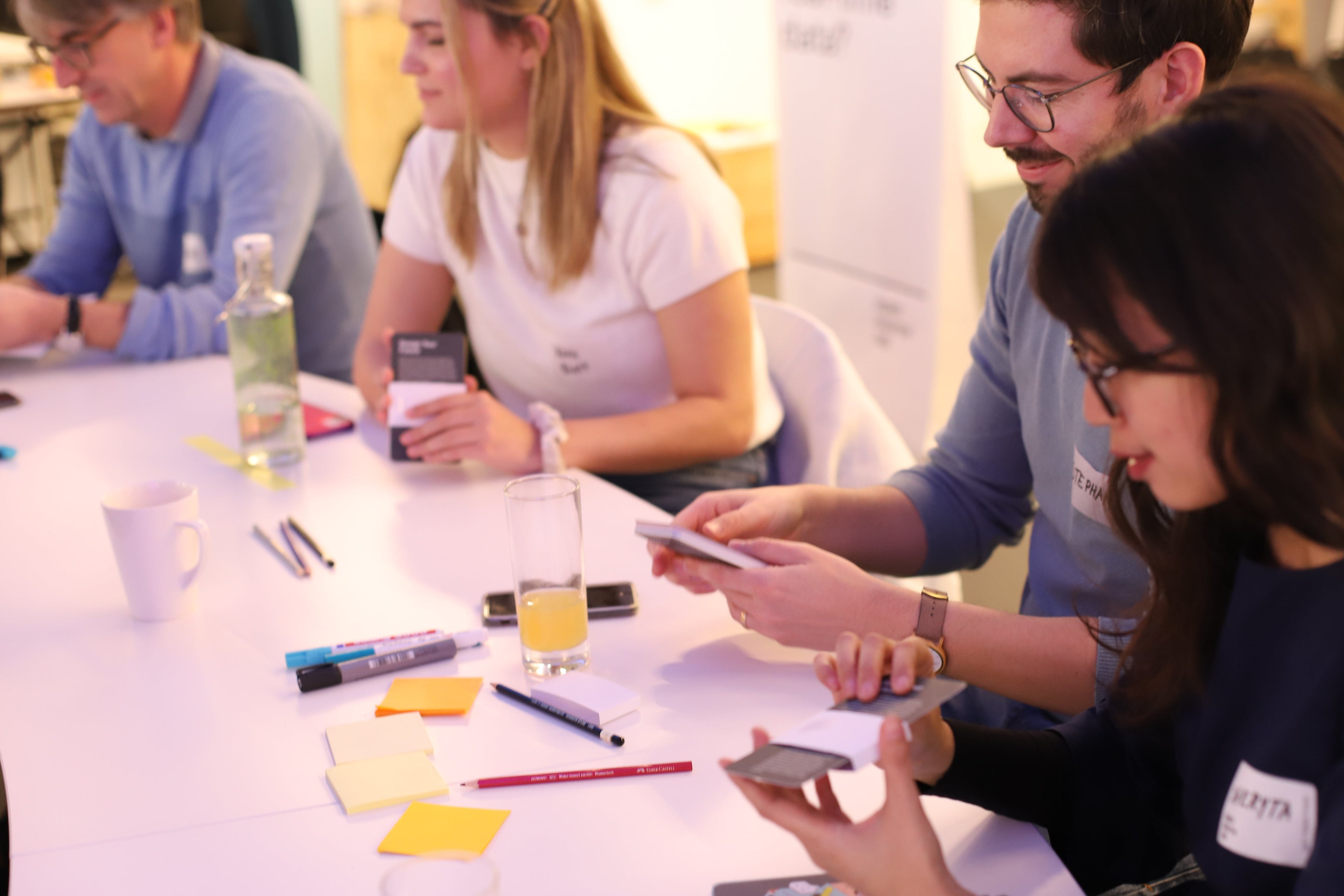 A group of people collaborating around a white table, holding smartphones and interacting in a bright indoor office or meeting space.